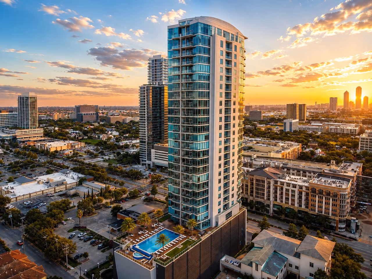 Aerial photograph of 2727 Kirby in low golden light: a curved glass tower with balconies rises at the centre of the frame above a dark podium whose roof terrace holds a rectangular blue pool, a circular spa, rows of white loungers and palm trees. Two glass high-rises stand behind it to the left, a low-rise brick and stucco block with arched windows to the right, shopping-center roofs and parking lots fill the foreground, and Houston mid-rise and high-rise buildings sit on the horizon under gold-lit clouds with the sun low at the far right.